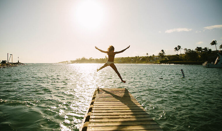 A person joyfully leaps from a dock into the water, illuminated by a bright sun with a serene landscape in the background.