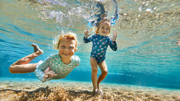 Two children underwater in a swimming pool, both smiling and wearing swimsuits. They appear to be enjoying the experience.