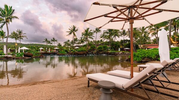 A tropical resort pool area with lounge chairs, umbrellas, palm trees, and a calm water body reflecting a pink sunset sky.