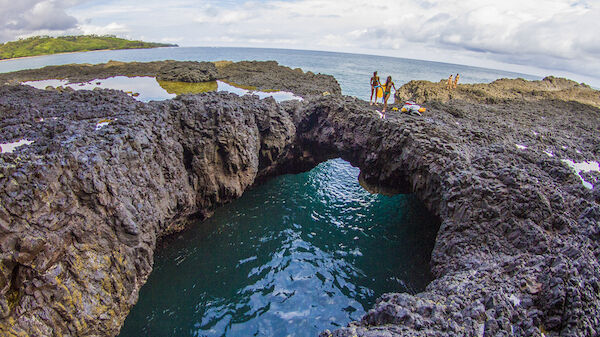 A natural rock formation with a water-filled hole is seen, surrounded by rocky terrain, with a few people in the background near the ocean.