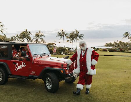 A person in a Santa outfit stands next to a red Jeep labeled "Santa," with palm trees and green grass in the background.
