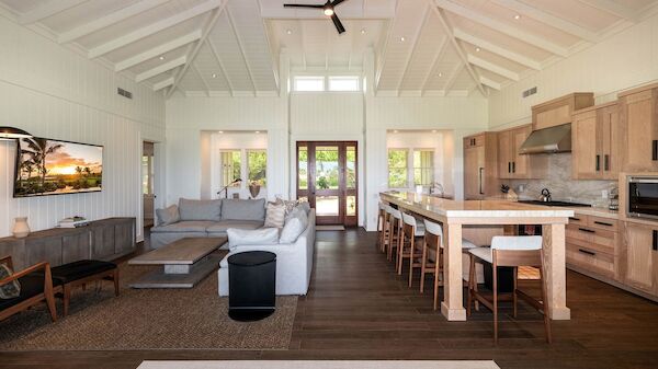 A bright, open-plan living and kitchen area with a vaulted ceiling, white walls, a large sofa, island/bar seating, and wooden cabinetry.