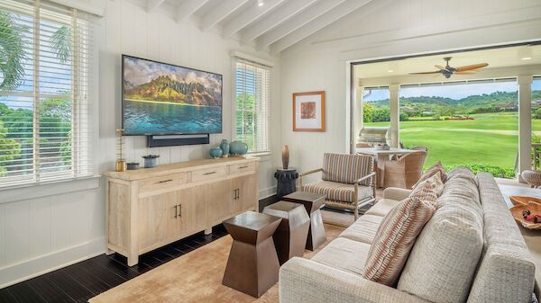 Bright living room with a wall TV, white paneled ceiling, beige sofa, wooden coffee tables, and a view of green golf course through large windows.