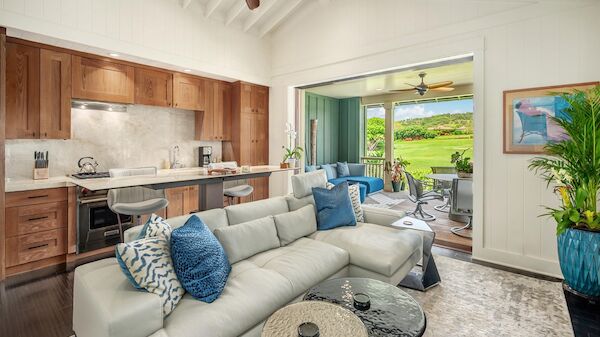 A modern open-plan living area with a cream sofa, blue throw pillows, and a kitchen featuring wooden cabinets; a leafy green view through a glass door.