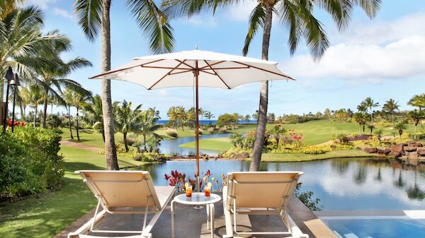 Two lounge chairs under a parasol by a pool, with palm trees and a serene lake/green landscape beyond, sunny tropical resort vibe.