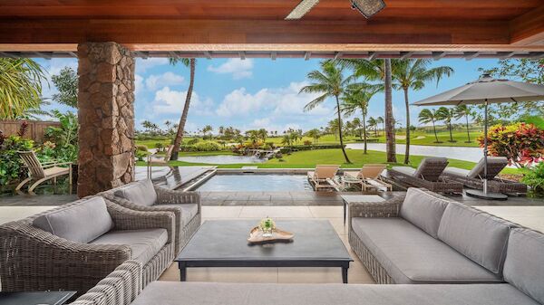 A luxurious outdoor lounge with gray cushioned sofas around a coffee table, a manicured lawn, palm trees, and a pool or view of the ocean in the background.