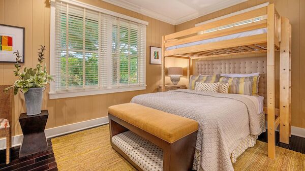 A cozy bedroom with a wooden bunk bed over a full-size bed, tufted bench at the foot, beige walls, white blinds, and a potted plant by the window.