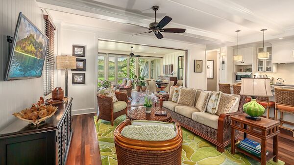Cozy living area with wicker and wooden furniture, a green rug, wall-mounted TV, and an open kitchen in the background, bright and inviting.