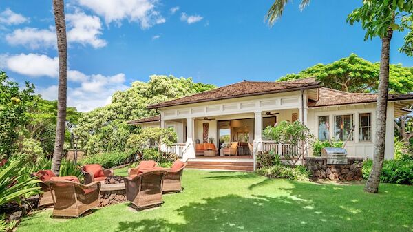A tropical bungalow with a spacious grassy yard, palm trees, and wicker seating on a patio under a bright blue sky.