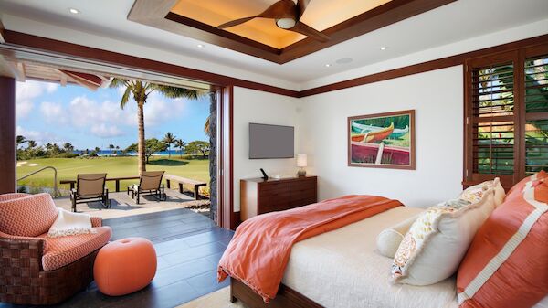A bright hotel room with a large bed, orange accents, a sitting area, and a view of a lush patio and palm trees outside.