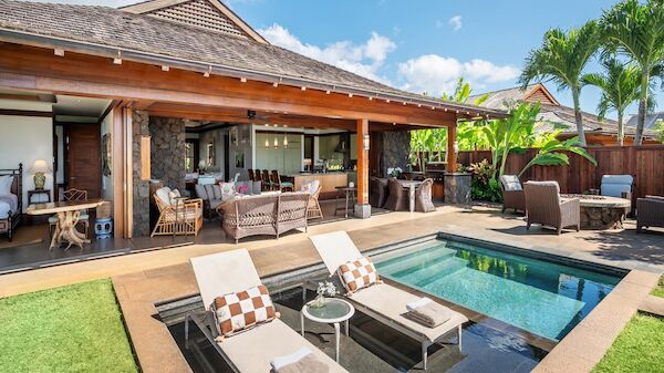 A luxurious backyard with a covered patio, seating area, a pool and hot tub, lounge chairs, and tropical landscaping under a sunny sky.