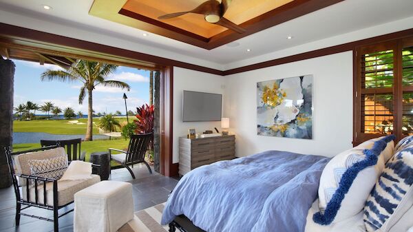 A luxurious tropical bedroom opens to an outdoor patio with palm trees and a lawn, featuring a blue bed, white furniture, and ocean views.