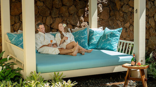 Two people relax on a blue-cushioned outdoor daybed under a shaded pergola, wearing robes, with tropical plants and a small side table nearby.