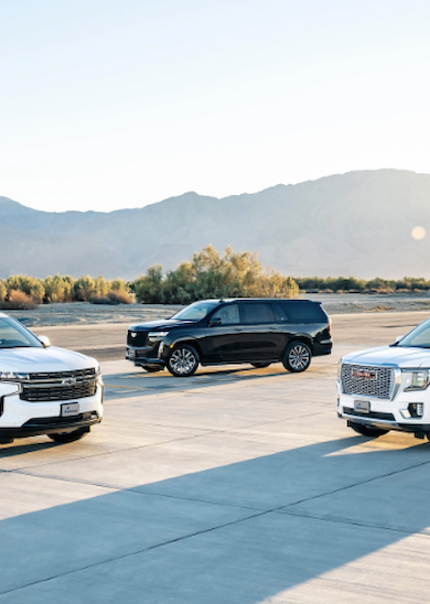 The image features three SUVs parked in an open area, with mountains in the background under a clear sky.
