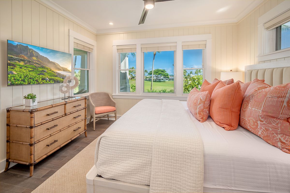 A bright, modern bedroom with a white bed, coral-pink pillows, a wooden dresser with a TV, a small chair, and large windows showing green scenery, ending the sentence.