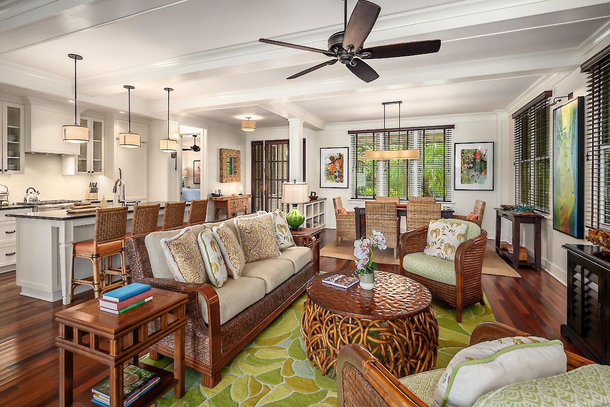 A bright open-concept living area with a kitchen bar, ceiling fan, wicker seating, green rug, and large windows letting in natural light.