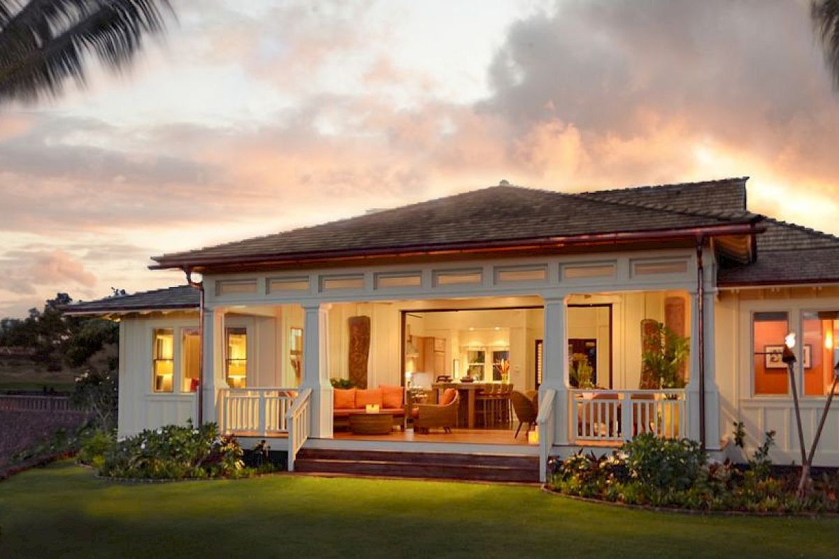 A cozy single-story house with a bright open porch, warm interior lights, lush lawn, palm trees, and a sunset sky behind it.