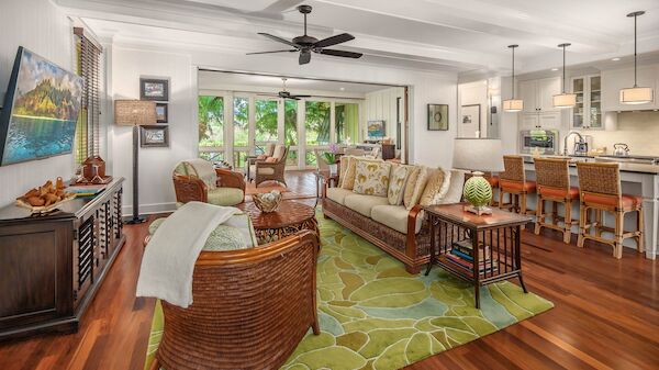 A bright, open living-dining area with wicker seating, a green patterned rug, hardwood floors, large windows, and a modern kitchen bar&mdash;cozy and airy.