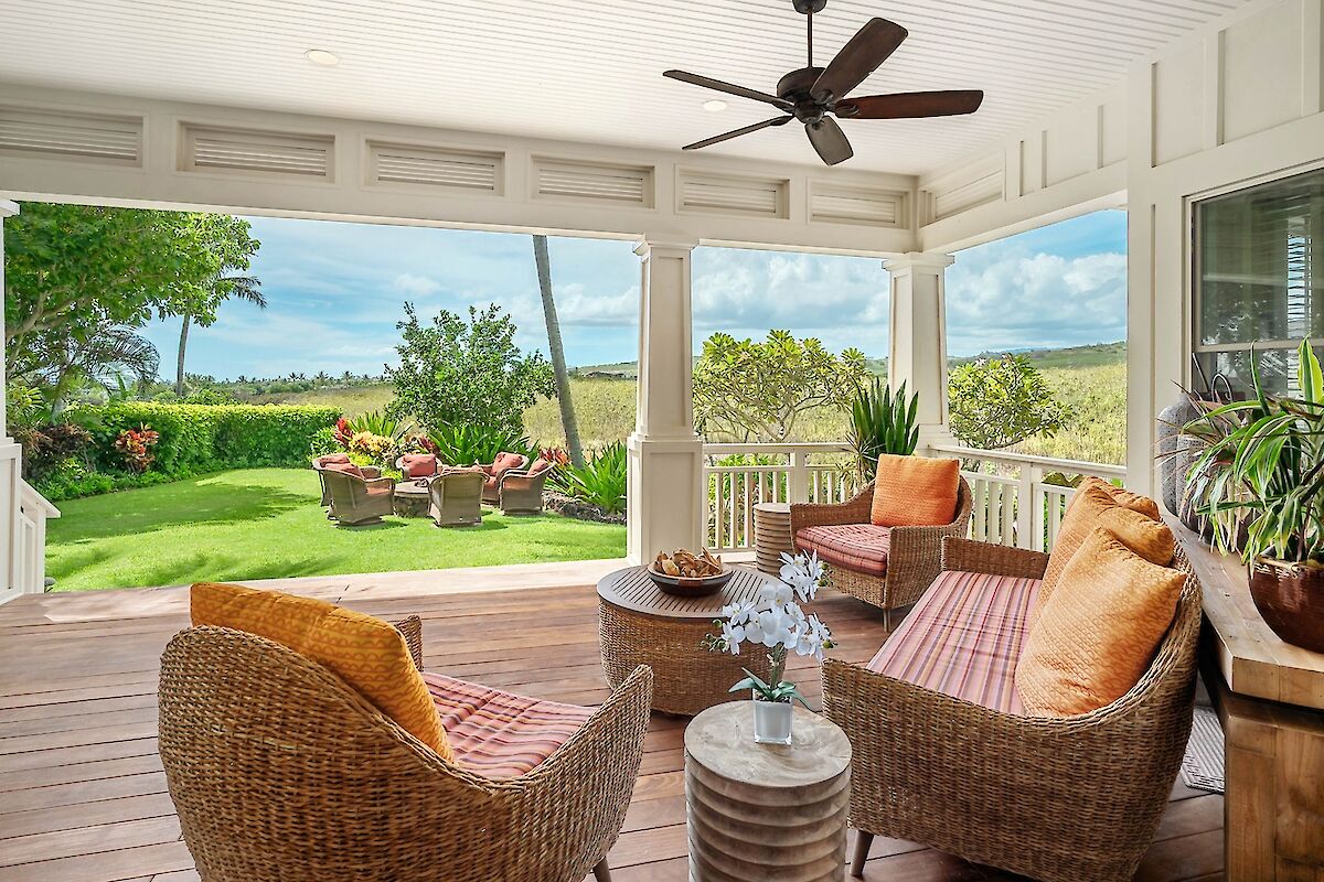 A sunny screened porch with wicker furniture, orange cushions, ceiling fan, and a view of a lush green lawn and ocean beyond.