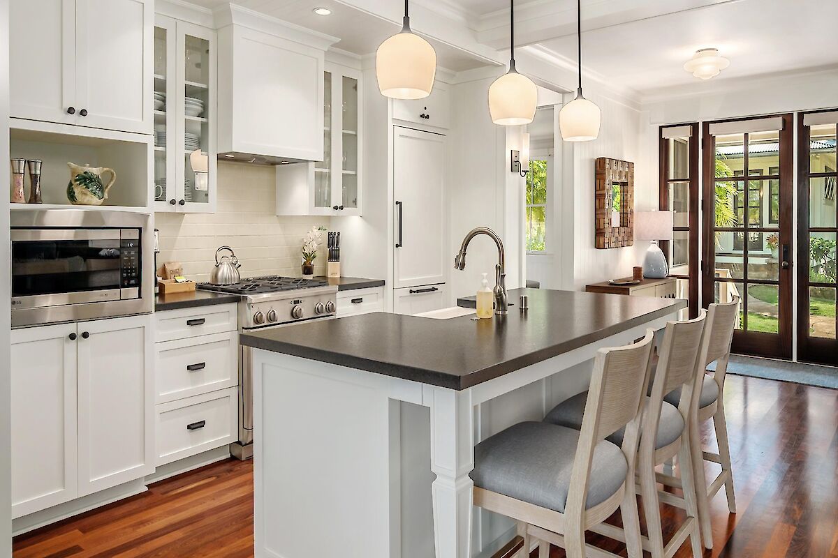 A bright, modern kitchen with white cabinets, a dark countertop island, pendant lights, stainless appliances, and a glass door leading outside.