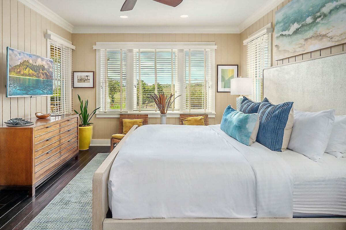 A bright, airy hotel-like bedroom with a white bed, blue pillows, wooden dresser, plants, and large windows letting in natural light.