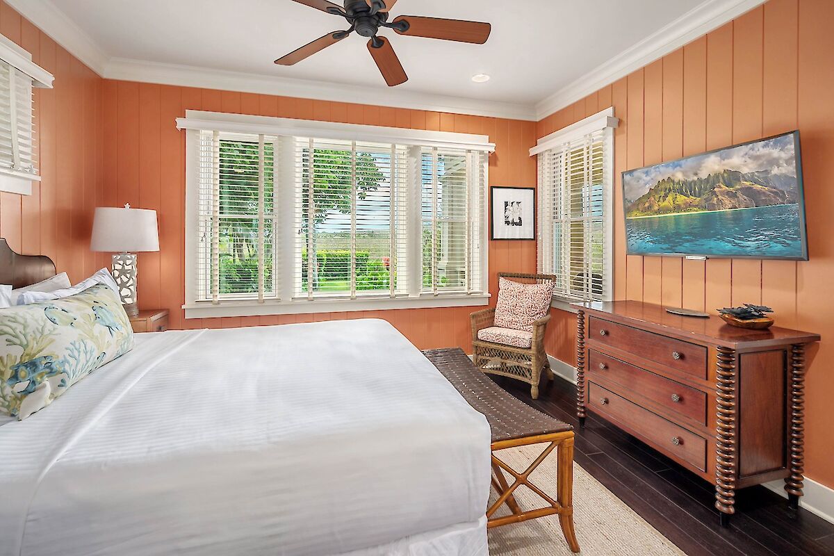 A cozy, sunny bedroom with burnt-orange walls, a large bed, white linens, wooden dresser, wicker chair, ceiling fan, and a panoramic window view, ending here.