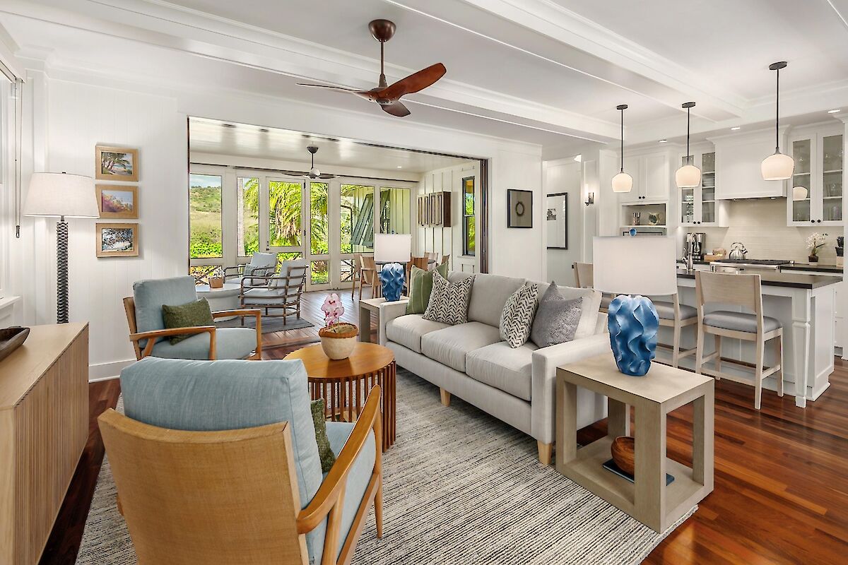 Cozy, bright living area with a light gray sofa, blue accent chairs, wooden floors, and an open kitchen with island seating, plus a ceiling fan.