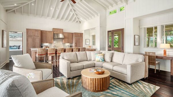 A bright, open living area with white walls, vaulted ceiling, and ceiling fan; a tan sectional, round coffee table, and a kitchen island in the background.