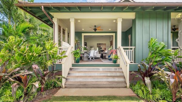 A bright, inviting tropical house with a shaded porch, lush plants, and outdoor seating, perfect for relaxing by the stairs.