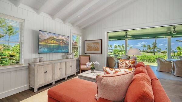A bright coastal living room with a coral sofa, white paneled walls, ocean view through large windows, and tropical decor.