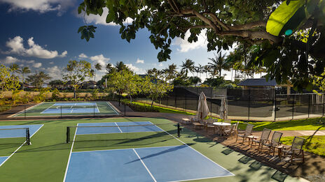A sunny outdoor tennis and pickleball court area with multiple courts, shaded seating, and palm trees in a tropical setting.