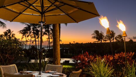 A tropical outdoor dining setup at sunset with a yellow umbrella, tiki torches, wicker chairs, and lush plants.