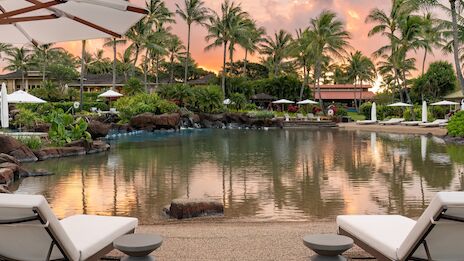 A luxurious resort poolside at sunset with palm trees, lounge chairs, and calm water reflecting the warm sky.