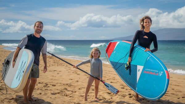 Three people on a sunny beach with two adults carrying surfboards and a child holding a paddle; ocean waves in the background.