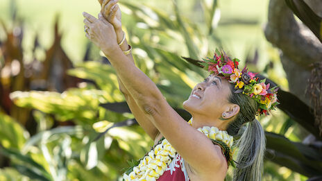 A performer wearing a flower lei and crown, smiling as she dances or blesses with hands raised, outdoors among tropical plants.