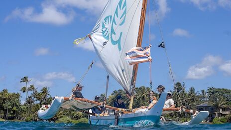 A small sailboat with a blue hull and a large white sail, on clear water near a tropical shoreline. End.