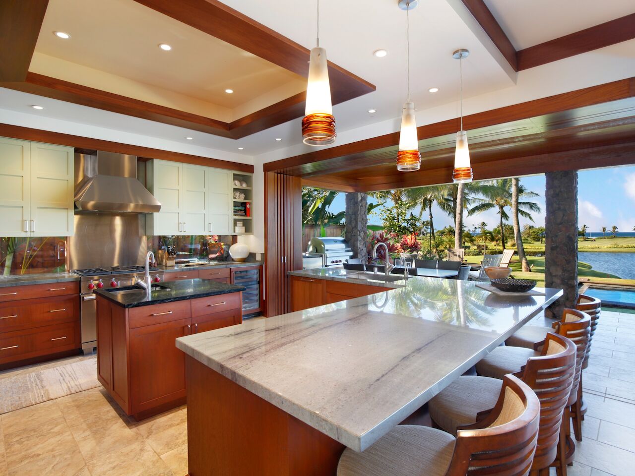 A modern outdoor-indoor kitchen with a central island, stainless steel appliances, pendant lights, and seating overlooking a sunny backyard lagoon.