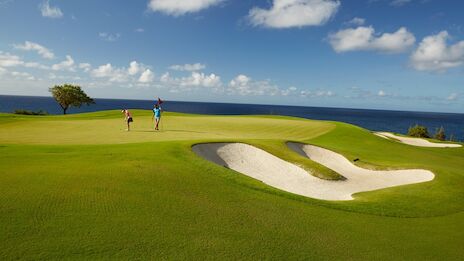 Two golfers walk on a lush golf course near the sea, with a sandy bunker and a bright blue sky scattered with clouds.