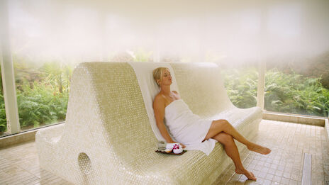 A woman in a white towel sits lounging on a curved, tiled bench inside a bright spa-like area with greenery outside, bare feet, and a small bouquet.