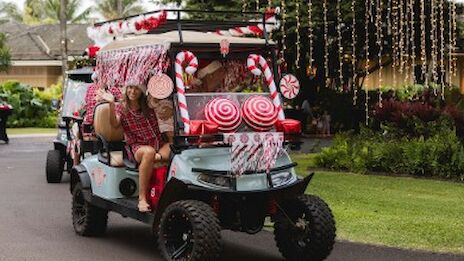 A festive golf cart decorated like a candy cane parade float with oversized peppermint swirls, red bows, and two riders smiling as they drive.