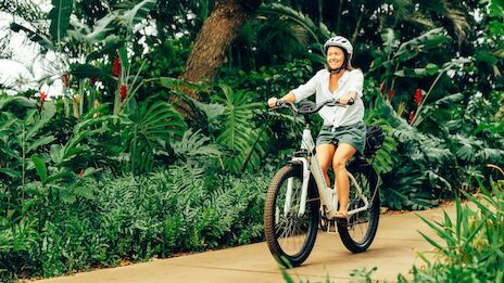 A person in a helmet rides a bike along a sandy path through lush tropical greenery.