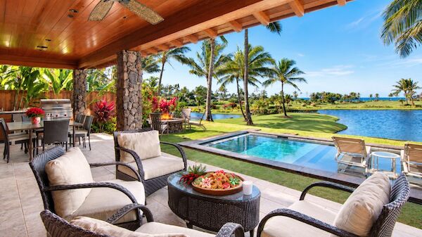 A tropical outdoor seating area overlooking a pool and lawn with palm trees and a sunny view of the water, perfect for lounging and dining.