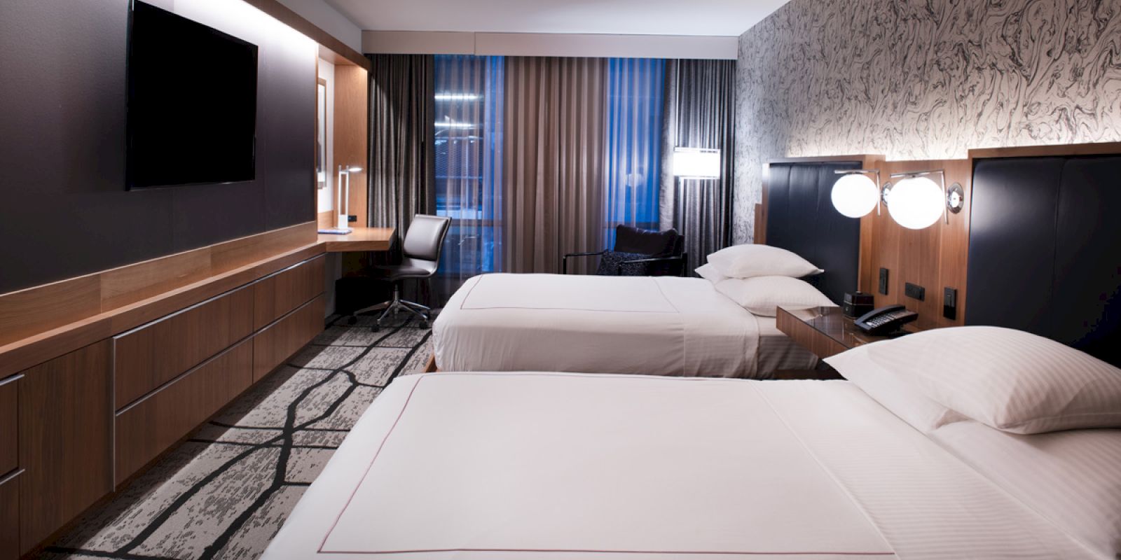 A modern hotel room with two beds, wall-mounted TV, a work desk near the window, and stylish lighting on the headboards, creating a cozy atmosphere.