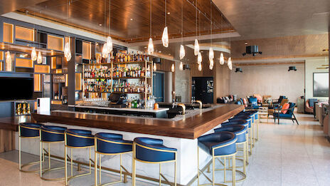 A modern bar with a large wooden counter, blue bar stools, hanging lights, and shelves filled with bottles, set in a stylish, open seating area.