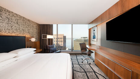 A modern hotel room featuring a large bed, a flat-screen TV, a desk with a chair, a seating area, and a window with a city view ending the sentence.