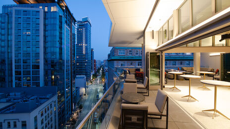 A modern urban balcony at twilight with seating and tables, overlooking a cityscape with tall buildings and illuminated streets.