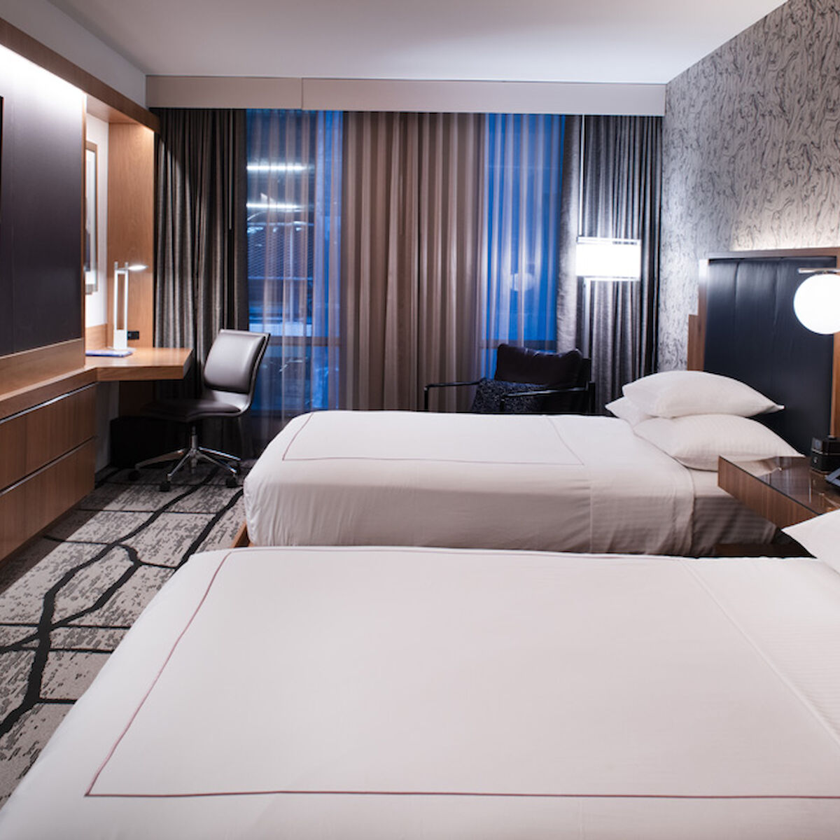 A modern hotel room featuring two neatly made beds, a desk with a chair, a large TV on the wall, and stylish lighting and decor.