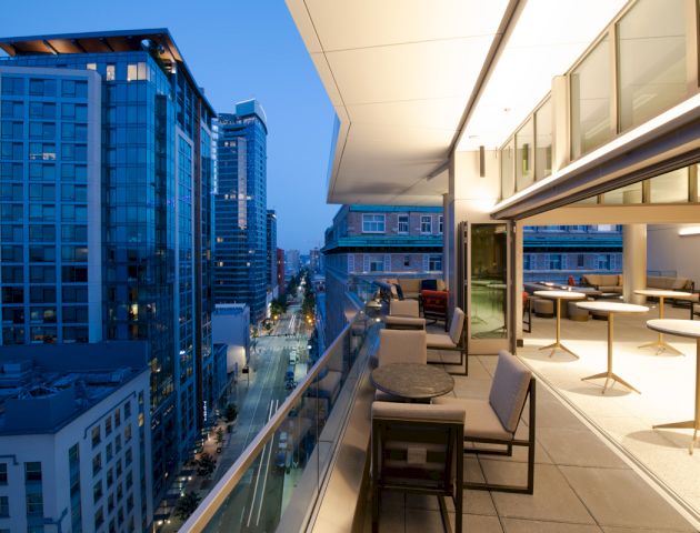 A modern cityscape at dusk is seen from a rooftop terrace with chairs, tables, and glass railings. Tall buildings line the well-lit street below.