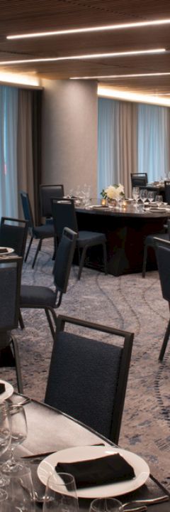 The image shows a decorated banquet hall with round tables set for an event, featuring black chairs, white plates, glasses, and flower centerpieces.