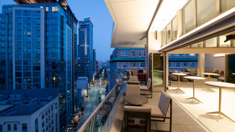 An outdoor terrace at dusk with a city view, featuring seating areas and tables. Modern buildings line the street below, creating a vibrant urban scene.